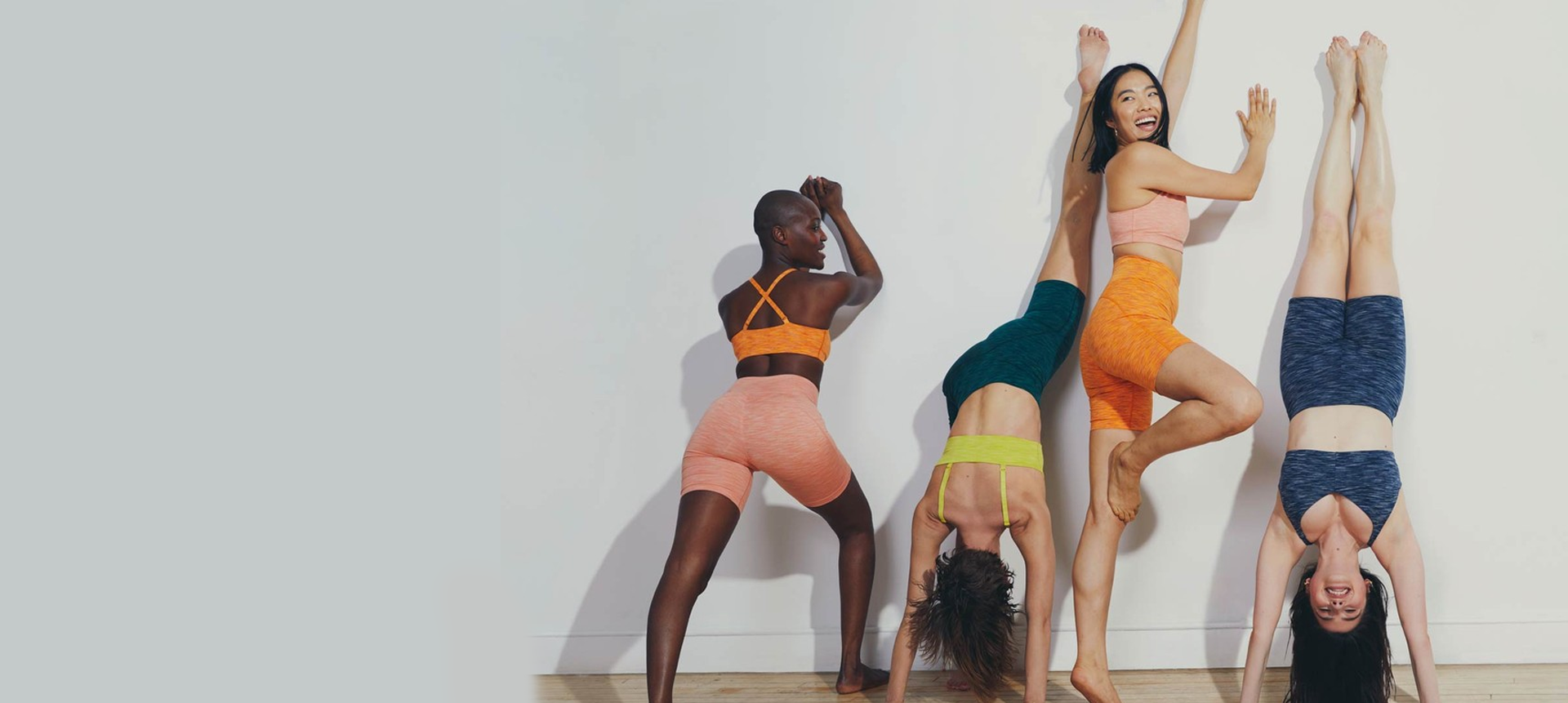 Four women in colorful workout outfits hanging from a bar against a white wall.